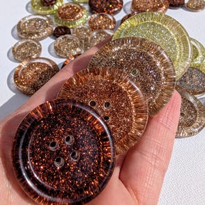 Puede incluir: Una colección de botones brillantes en varios tamaños y tonos de marrón, dorado y transparente. Los botones tienen cuatro agujeros y se sostienen en una mano, mostrando sus detalles brillantes. Botones para coser y manualidades.