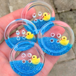 May include: Four clear, round buttons with a blue glitter base and a yellow rubber duck. The buttons have a bubble design and are held in a person's hand. The buttons are a craft or sewing supply.