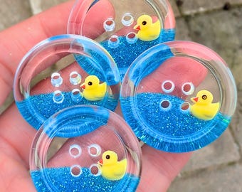 Op de afbeelding: Vier heldere, ronde knopen met een blauwe glitterbasis en een gele badeend. De knopen hebben een bubbelontwerp en worden in de hand van een persoon gehouden. Naai- of knutselbenodigdheden.