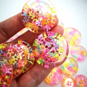 Op de afbeelding: Transparante, ronde knopen met kleurrijke stervormige confetti. De knopen hebben meerdere gaten om te naaien en een transparante rand. De confetti bevat tinten roze, geel, oranje en groen.