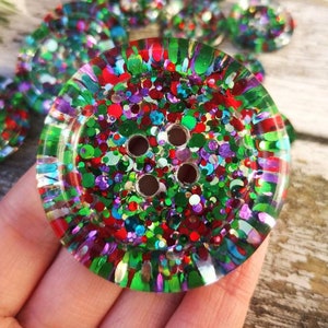 May include: A large, round, multi-colored button with a glittery finish. The button has four holes and is made of clear resin with embedded glitter in shades of red, green, blue, purple, and gold.