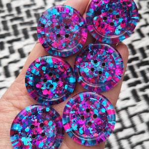May include: Six colourful buttons with a glittery finish. The buttons are blue, pink, and purple with a speckled pattern. Each button has four holes.