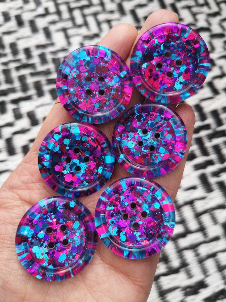 May include: Six colourful buttons with a glittery finish. The buttons are blue, pink, and purple with a speckled pattern. Each button has four holes.