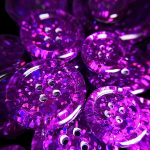 Peut inclure: Gros plan sur un tas de boutons en paillettes violettes. Les boutons sont ronds et ont quatre trous chacun. Les paillettes sont irisées et reflètent la lumière.