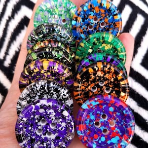 May include: A collection of ten colorful buttons with a speckled paint design. Each button has four holes and is made of clear resin. The buttons are arranged in a stack, with the largest button at the bottom and the smallest button at the top.