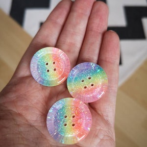 May include: Three rainbow iridescent buttons with four holes each. The buttons are made of a clear plastic with a rainbow glitter embedded in the plastic.
