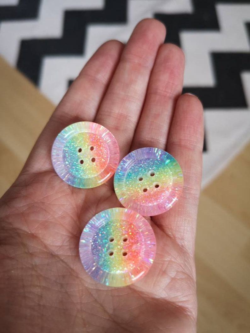 May include: Three rainbow iridescent buttons with four holes each. The buttons are made of a clear plastic with a rainbow glitter embedded in the plastic.