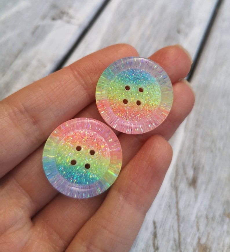 May include: Two iridescent rainbow glitter buttons with four holes each. The buttons are round and have a light pink, orange, yellow, green, and blue color gradient.