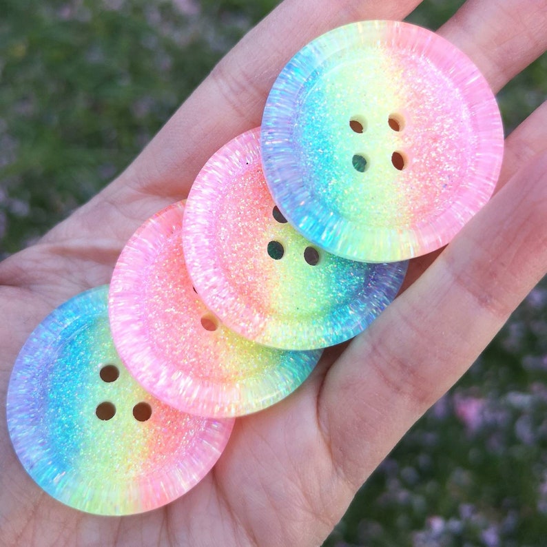 May include: Four iridescent rainbow buttons with a glittery finish and four holes each. The buttons are stacked on top of each other.