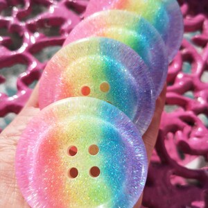 May include: Four rainbow glitter buttons with four holes each. The buttons are stacked on top of each other and are made of clear resin with a rainbow glitter embedded in the resin.