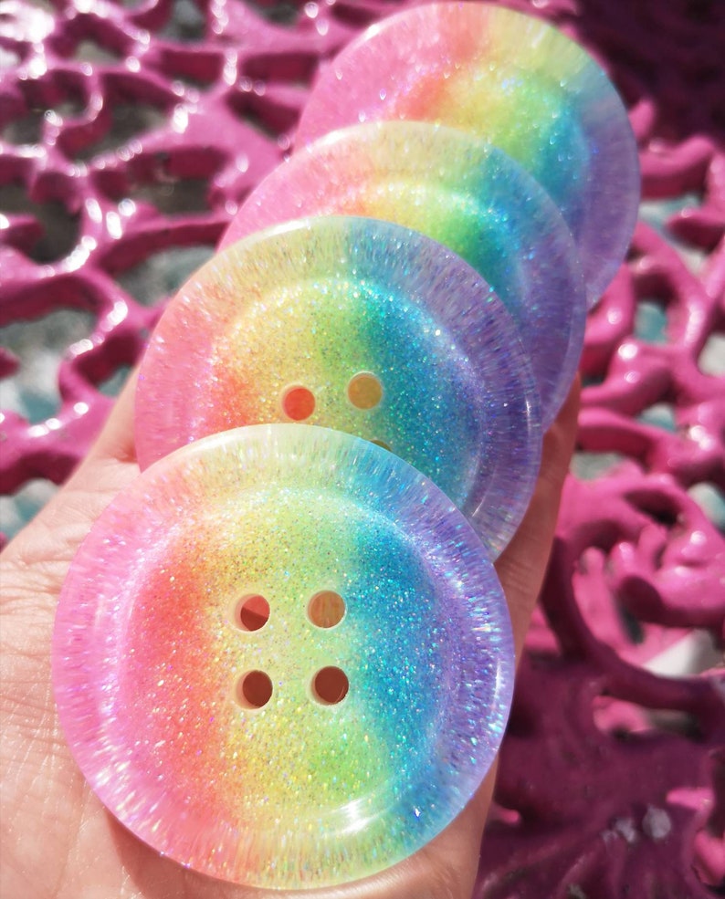 May include: Four rainbow glitter buttons with four holes each. The buttons are stacked on top of each other and are made of clear resin with a rainbow glitter embedded in the resin.