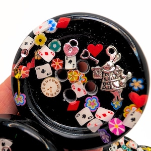 May include: Black resin coaster with embedded charms. The coaster features playing card suits, flowers, a clock face, a heart, a key, and a rabbit figure. The design is inspired by Alice in Wonderland.