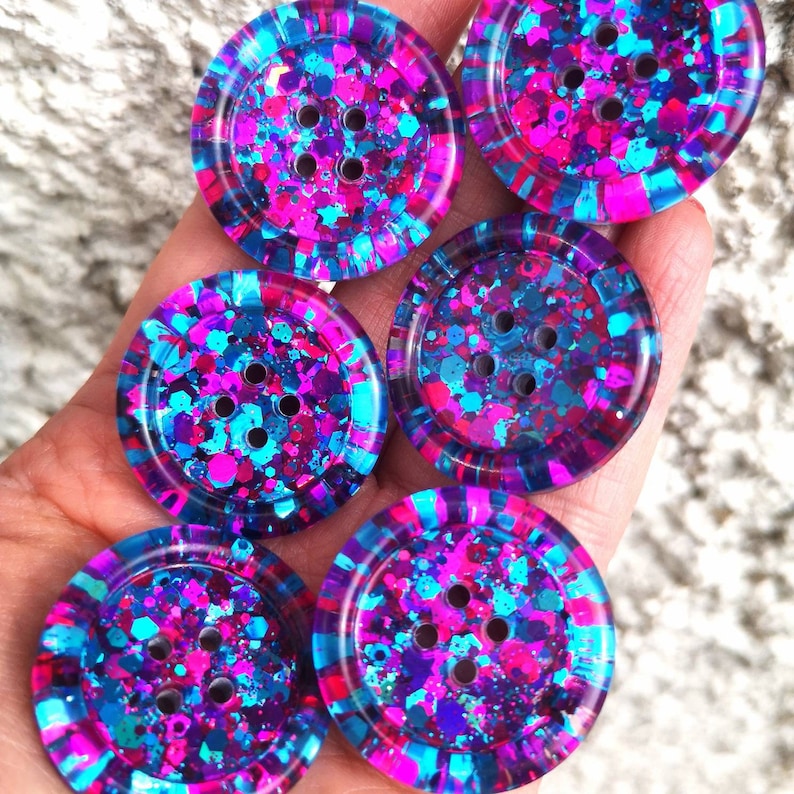 May include: Six round, colourful buttons with a glittery finish. The buttons are blue, pink, and purple with a white outline. Each button has five holes.