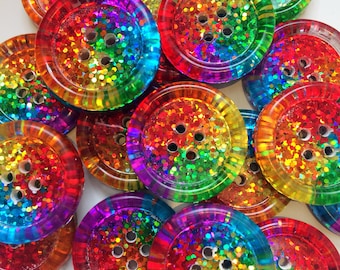 May include: A collection of rainbow glitter buttons with a multi-colored iridescent finish. The buttons have a raised rim and five holes in the center.