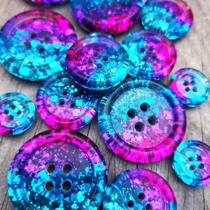 Könnte beinhalten: Eine Sammlung bunter Knöpfe mit einem blauen und rosa Ombre-Effekt und Glitzer, der in das Harz eingebettet ist. Die Knöpfe sind in verschiedenen Größen und haben jeweils vier Löcher.