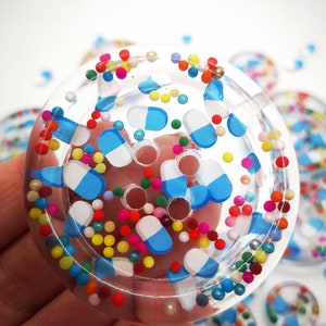 Puede incluir: Un botón de resina transparente con un diseño de chispas de colores y formas de píldoras azules y blancas. El botón tiene cuatro agujeros para coser.