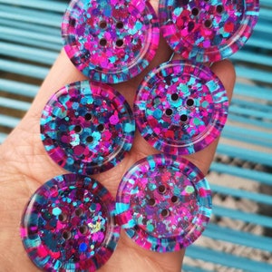 May include: Six round, colourful buttons with a glittery finish. The buttons are pink, blue, and purple with a mix of glitter colours. Each button has five holes.