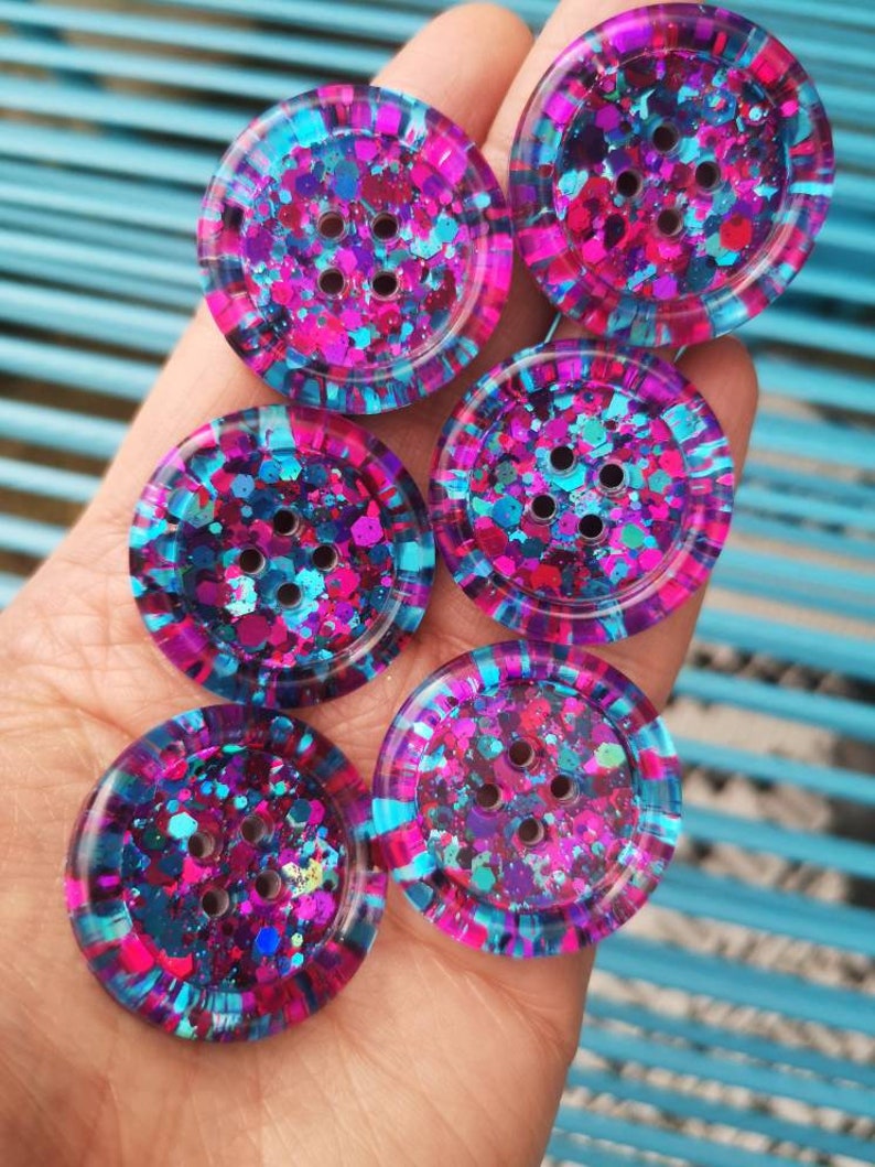 May include: Six round, colourful buttons with a glittery finish. The buttons are pink, blue, and purple with a mix of glitter colours. Each button has five holes.