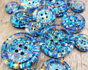 May include: Assortment of blue and gold glitter resin buttons with four holes. The buttons are various sizes and have a clear resin coating.