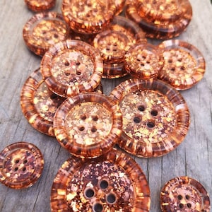 Könnte beinhalten: Eine Sammlung von 14 kupferfarbenen Knöpfen mit glitzerndem Finish. Die Knöpfe sind rund und haben jeweils vier Löcher. Sie sind auf einer Holzoberfläche zu einem Stapel angeordnet.