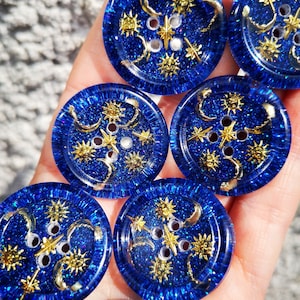 Könnte beinhalten: Nahaufnahme von mehreren runden, blauen Knöpfen mit glitzerndem Finish. Jeder Knopf hat goldfarbene Himmelsdesigns, darunter Monde, Sterne und Sonnen. Die Knöpfe haben vier Löcher zum Nähen und eignen sich für verschiedene Bastelprojekte.