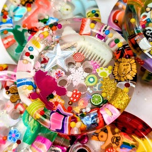 Op de afbeelding: Een verzameling kleurrijke hars onderzetters met ingebedde decoratieve elementen. Elke onderzetter heeft een heldere harsbasis gevuld met diverse kleine objecten, waaronder glitter, sterren, fruitschijfjes en miniatuurbeeldjes. De onderzetters hebben een glanzende afwerking.