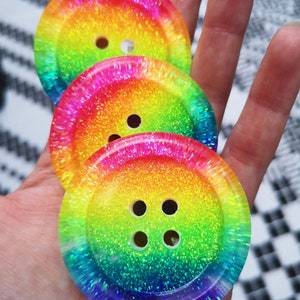 Puede incluir: Tres botones de purpurina arcoíris con un efecto de capas. Los botones tienen un degradado arcoíris con un acabado brillante. El botón más grande tiene cuatro agujeros, el botón del medio tiene dos agujeros y el botón más pequeño tiene un agujero.