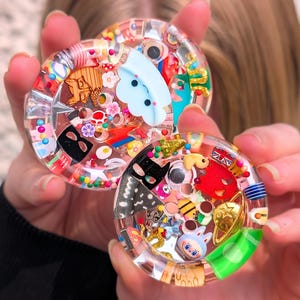 Könnte beinhalten: Drei transparente Harz-Untersetzer, gefüllt mit bunten Miniatur-Objekten. Jeder Untersetzer hat eine einzigartige Ansammlung von kleinen Spielzeugen, Perlen und Charms, darunter eine Wolke, eine Maske und eine Rakete. Die Untersetzer werden von einer Person gehalten.