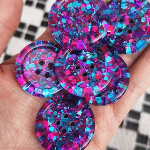 May include: Five colourful buttons with a glittery finish. The buttons are round and have four holes each. The buttons are a mix of pink, blue, and purple.