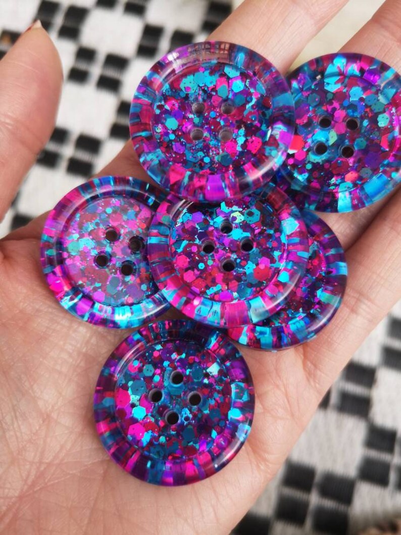 May include: Five colourful buttons with a glittery finish. The buttons are round and have four holes each. The buttons are a mix of pink, blue, and purple.