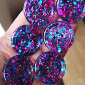 May include: Six round, colourful buttons with a glittery finish. The buttons are a mix of pink, blue, and purple with a clear resin coating. Each button has four holes for sewing.