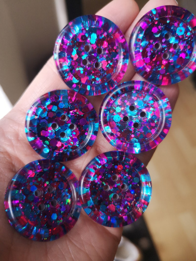 May include: Six round, colourful buttons with a glittery finish. The buttons are a mix of pink, blue, and purple with a clear resin coating. Each button has four holes for sewing.