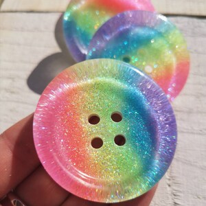 May include: Four iridescent rainbow glitter buttons with four holes each. The buttons are made of clear resin with a rainbow glitter embedded in the resin. The buttons are arranged in a stack with the largest button on the bottom and the smallest button on top.
