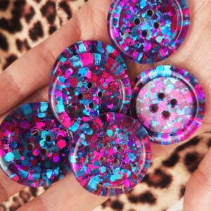 May include: Five round, colourful buttons with a glittery finish. The buttons are pink, blue, and purple with a black background. Each button has four holes.