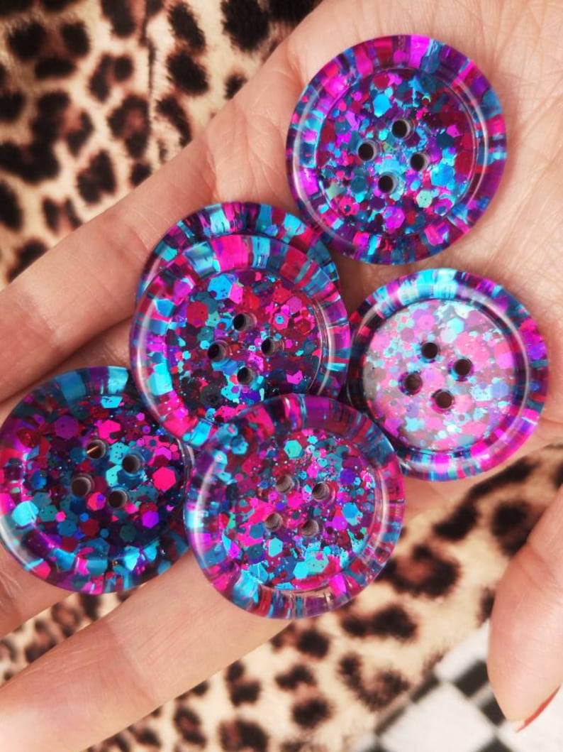 May include: Five round, colourful buttons with a glittery finish. The buttons are pink, blue, and purple with a black background. Each button has four holes.