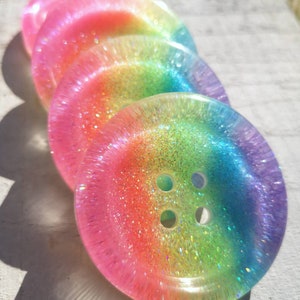 May include: Four iridescent rainbow buttons with glitter embedded in the resin. The buttons have four holes and are stacked on top of each other.