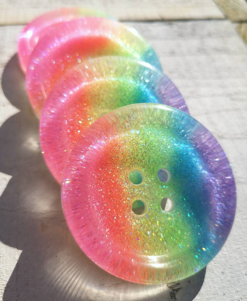 May include: Four iridescent rainbow buttons with glitter embedded in the resin. The buttons have four holes and are stacked on top of each other.