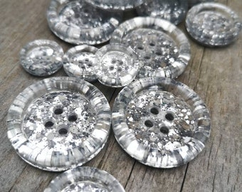 May include: A pile of silver glitter buttons of various sizes. The buttons have a clear plastic base with silver glitter embedded in the plastic. The buttons have four holes for sewing.