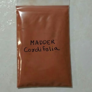 May include: A transparent plastic bag containing a reddish-brown powder. The bag is labelled with the handwritten text "Madder Cordifolia". The powder has a fine consistency, possibly for use as a dye or pigment.
