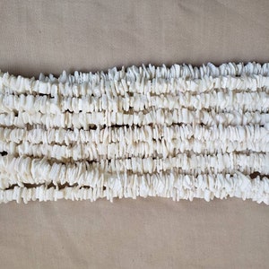 Puede incluir: Primer plano de varios hilos de cuentas de virutas blancas. Las cuentas tienen forma irregular y parecen estar hechas de un material natural. El fondo es de color beige neutro.