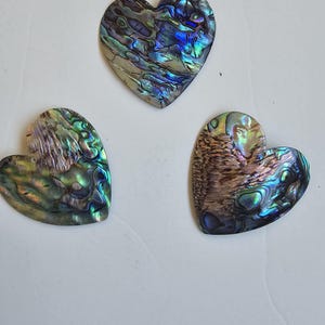 Könnte beinhalten: Drei regenbogenfarbene Abalone-Muschel-Herzformen in Blautönen, Grüntönen und Braun.