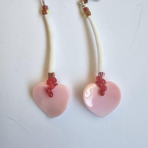 May include: A pair of dangle earrings featuring pink heart-shaped pendants. The earrings have white, curved stems with red and gold beads near the top and bottom, and silver-colored ear wires. The earrings are displayed on a white surface.