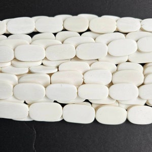 May include: A close-up of a strand of white oval-shaped beads. The beads are smooth and polished, and they are arranged in a staggered pattern.