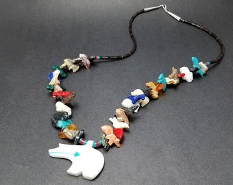 May include: A Native American-style necklace with a white carved bear pendant. The necklace is made of black cord and features a variety of colorful carved animal figures.