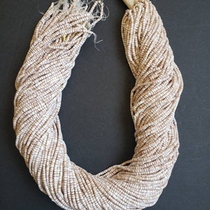 May include: A large bundle of small, off-white and brown beads, tightly strung together. The beads are cylindrical and arranged in a U-shape against a dark background. The ends of the strands are tied and wrapped.