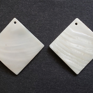 Puede incluir: Dos piezas de concha blancas en forma de diamante con un agujero en la parte superior para colgar. Las piezas de concha tienen un brillo natural e iridiscente.