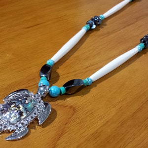 May include: A silver turtle pendant necklace with a mother of pearl shell inlay. The necklace is made with white bone beads, black beads, and turquoise beads.