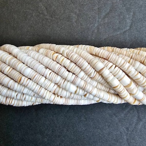 May include: A strand of white and cream-colored shell beads. The beads are round and flat, and they are arranged in a loose, flowing pattern.