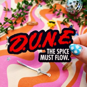 May include: A black sticker with the word "DUNE" in red, and the text "THE SPICE MUST FLOW." A miniature silver sword is in the foreground. The background has a retro, colorful pattern with a few small decorative items.