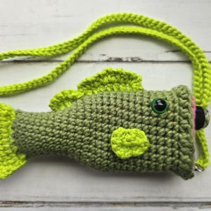 May include: A crocheted green and yellow bass fish purse with a black button eye and a pink and black detail on the side. The purse has a green and yellow crocheted strap.